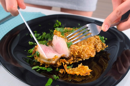 Woman hands cutting breaded chicken breast cutlet serving with greenery on black plate. Tasting cooked golden-brown crispy chicken poultry fillet. Culinary cuisine recipe, prepare food dish.