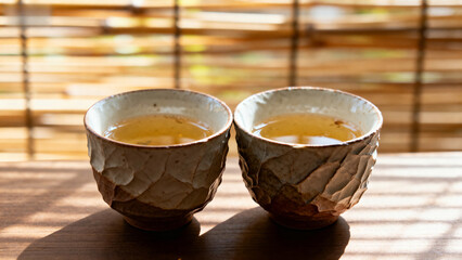 Two ceramic tea cups filled with tea on a wooden table, illuminated by natural light through bamboo blinds