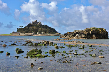 Saint Michael's Mount in Cornwall at high tide with submerged causeway, mossy green rocks, and castle in the background under blue summer sky