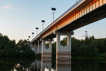 Golden sunlight casts a warm hue on the contemporary bridge, while tranquil waters mirror its beauty amidst lush greenery