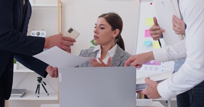 Very busy business woman sitting at the desk with notebook computer, surrounded by her colleagues, and having too much duties things to do. Business concept of hard working and multitasking.