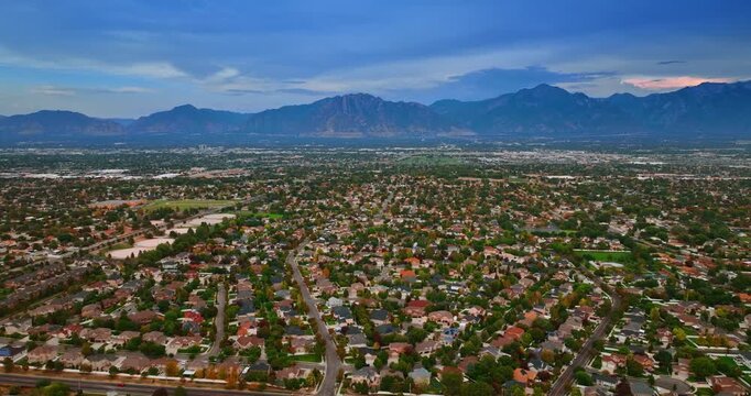 Vast green low-rise residential urban landscape. Mountain range silhouette at backdrop. Drone flight above Salt Lake City, Utah, USA at sunset.