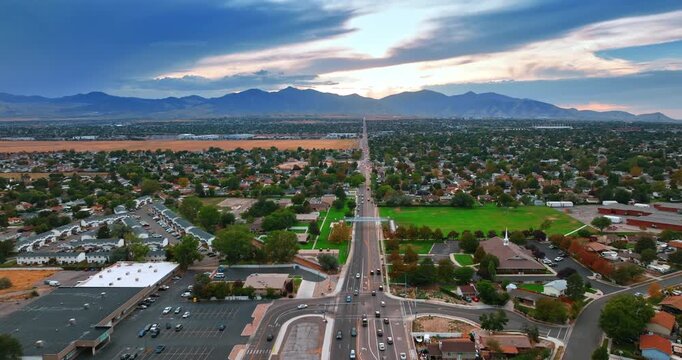 Transport goes by the long straight highway crossing the green neighborhoods. Mountain range silhouette at backdrop. Salt Lake City, Utah, USA at sunset. Aerial view.