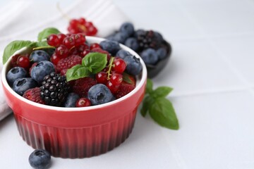 Different ripe berries and basil leaves in bowl on white tiled table, closeup. Space for text