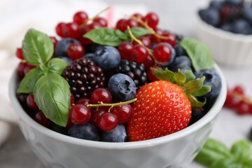 Different ripe berries and basil leaves in bowl on light table, closeup