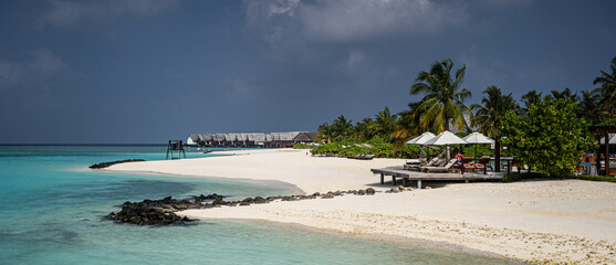White sandy beach on a Maldivian island with turquoise water, over water villas in the distance, palm trees, sun loungers with umbrellas, and volcanic rocks under a dramatic tropical sky.