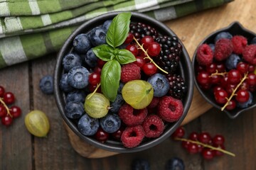 Different ripe juicy berries and basil in bowl on wooden table, flat lay
