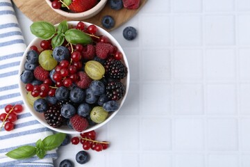 Different ripe juicy berries and basil in bowl on white tiled table, flat lay. Space for text