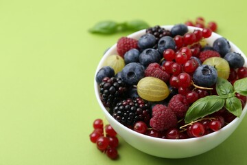Different ripe juicy berries and basil in bowl on green background, closeup. Space for text