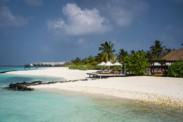 White sandy beach on a Maldivian island with turquoise water, over water villas in the distance, palm trees, sun loungers with umbrellas, and volcanic rocks under a dramatic tropical sky.