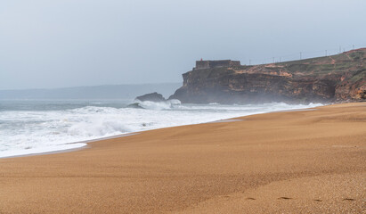 Waves crash onto a wide, empty sandy beach under a cloudy sky. A rugged cliff topped with an old fort overlooks the turbulent sea. The scene captures the raw, untouched beauty of a coastal landscape.