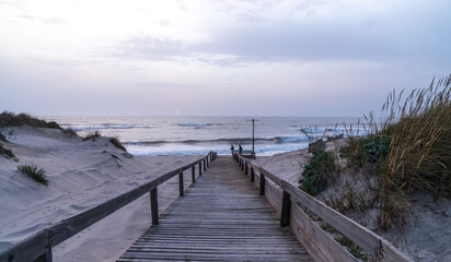 Naklejka premium A serene beach scene in Portugal. A wooden boardwalk through sand dunes to the ocean, where waves lap the shore under a cloudy sky. Sparse vegetation and evening light create a tranquil atmosphere.