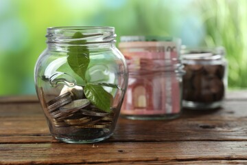 Glass jar with coins and sprout on wooden table against blurred background, closeup