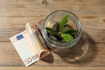 Glass jar with coins, sprout and banknotes on wooden table, top view