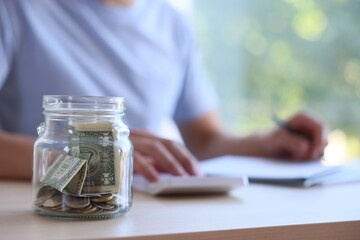 Fototapeta premium Glass jar with money and man budgeting at wooden desk, selective focus