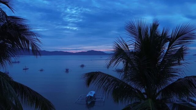 Sideways aerial glide beside palms in Port Barton San Vicente Palawan with calm bay moored outrigger boats distant islets blue hour sky and soft reflections