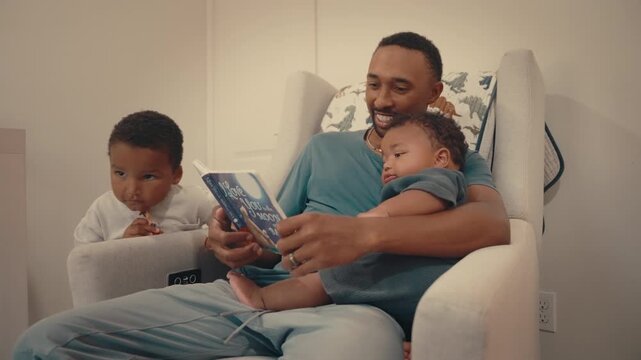Black dad reads to infant in rocking chair while toddler stands close. Brothers share bedtime routine, warm family bonding moment in cozy nursery