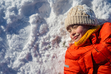 Child lying in fresh snow wearing bright winter clothes, enjoying sunny winter day