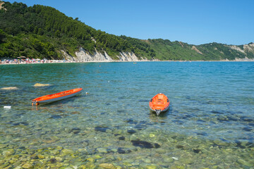 Sunny day at the beach across the cliffs in Portonovo Beach, Marche, Italy. Two red boats on the waves across the cliffs and sea