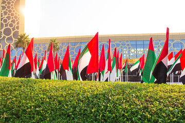 UAE flags waving in the wind in front of a modern glass building with geometric patterns, green hedge and palm trees under bright sunlight. Holiday, state event