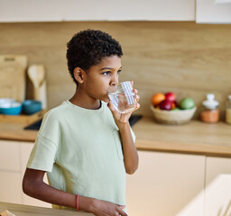 Portrait of a child, teen black boy,  drinks fresh Water in the Kitchen, holds a glass of pure...