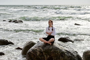 Young white woman with braids sits cross-legged on rocky shore. Overcast day with waves crashing, evoking tranquility and focus. Serene ocean backdrop enhances meditation scene