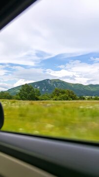 Car view shows a green meadow, treeline, & a blue sky-cloud topped mountain range