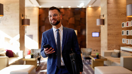 Smiling businessman in suit holding smartphone and briefcase walking through modern hotel lobby, successful corporate travel