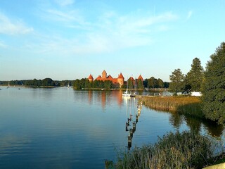 Panoramic view of Trakai Castle in Lithuania, a medieval red brick castle, situated on an island surrounded by a serene lake. The image captures the castle's towers and walls reflecting on the calm wa