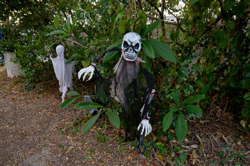 Tattered black-cloaked skull figure with chains stands among bushes and leaves. Spooky yard decoration scene with ghosts and foliage in daylight.