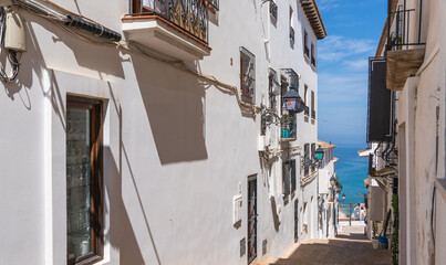 Charming narrow street in a coastal town with white traditional buildings, balconies, and a glimpse of the sea under a bright blue sky.