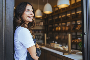 Bright Smiling Woman Stands At Bakery Entrance Holding Book, Wearing Apron, Surrounded By Fresh...