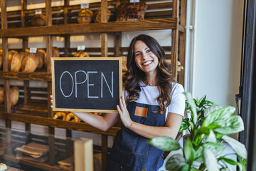 Smiling Bakery Employee Holds Open Sign In Front Of Fresh Bread Display