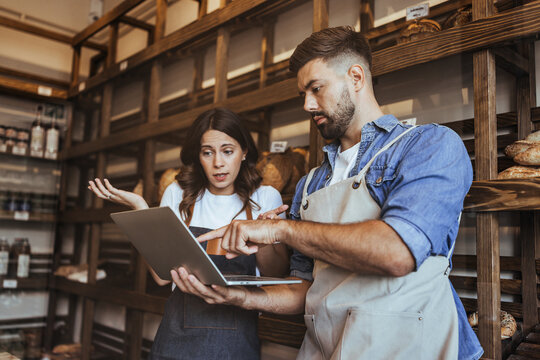Two Bakery Staff Collaborate On Laptop Troubleshooting And Planning In Rustic Store Setting