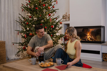 Couple Enjoying Coffee Together by the Christmas Tree in Decorated Room with Fireplace