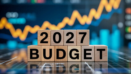 Wooden blocks spelling 2027 budget in front of blurred stock market graph with rising yellow and blue trend lines symbolizing financial planning, government spending, investment strategy, and economic