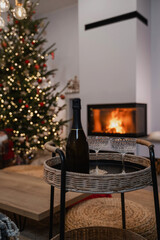 Champagne and glasses on a wicker tray in front of a decorated Christmas tree and fireplace