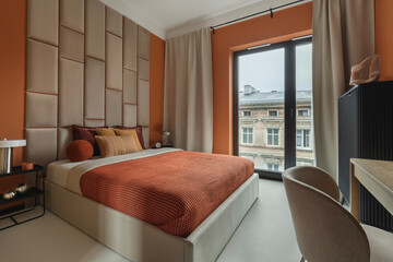 Modern bedroom with padded headboard, orange walls, and beige curtains, decorated with warm-toned pillows.