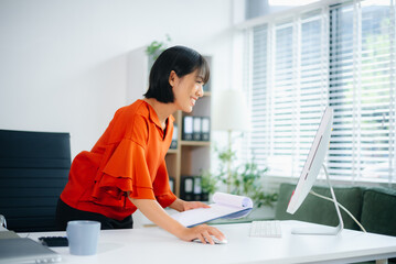 Happy Asian woman writes on clipboard while working on laptop at modern office desk. Concept of planning, multitasking, freelance work, and business productivity.