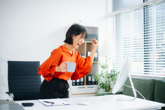 Smiling Asian woman raises papers in victory while celebrating success in a bright office. Concept of business win, joy, motivation, and achievement.
