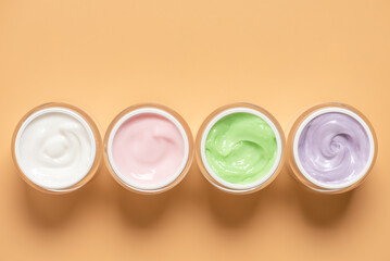 Various colored cosmetic creams in open jars on pastel yellow background in a row, top view, flat lay,