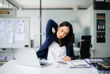 Asian businesswoman feeling neck pain from long office work, stretching at desk with laptop.