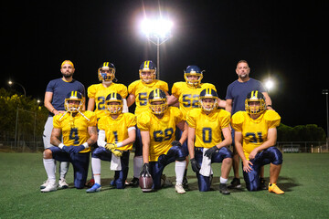 Fototapeta premium American football team and coaches standing and kneeling, posing on a green field at night