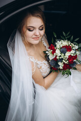 Naklejka premium Beautiful bride in lace wedding dress sitting in a car and holding bouquet of red, white and blue flowers. Smiling woman with veil looking out from vehicle on her wedding day. Elegant bridal portrait.