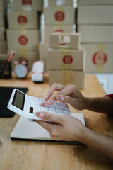 A person checking inventory with a tablet and notebook in a warehouse, surrounded by stacked cardboard boxes for shipping and logistics.