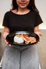 a person holding a black plate with Indonesian grilled chicken, steamed rice topped with fried shallots, sambal matah, and vegetable garnish.