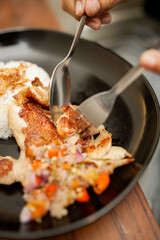 Close-up of Indonesian grilled chicken served with steamed rice, sambal matah, and vegetables on a black plate. A person eating traditional Asian food using fork and spoon.