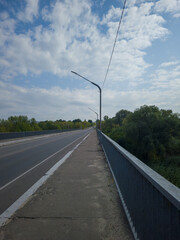 Long empty road stretches ahead, bordered by lush greenery and a clear blue sky, with streetlights lining the path, creating a serene and inviting atmosphere for travel