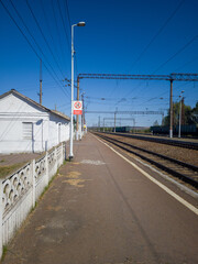 Train station platform with clear blue sky, featuring white building, railway tracks, and overhead lines, creating a serene travel atmosphere with open space