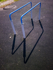 Blue and white outdoor exercise bars on a black rubber surface, casting long shadows in bright sunlight, showcasing fitness equipment in a public park environment
